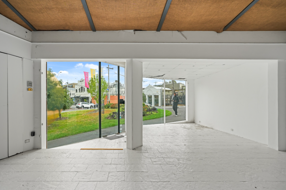 Bright, open pop-up shop interior with white walls, tiled flooring, large glass storefront windows, and a timber-lined ceiling overlooking a landscaped outdoor area.